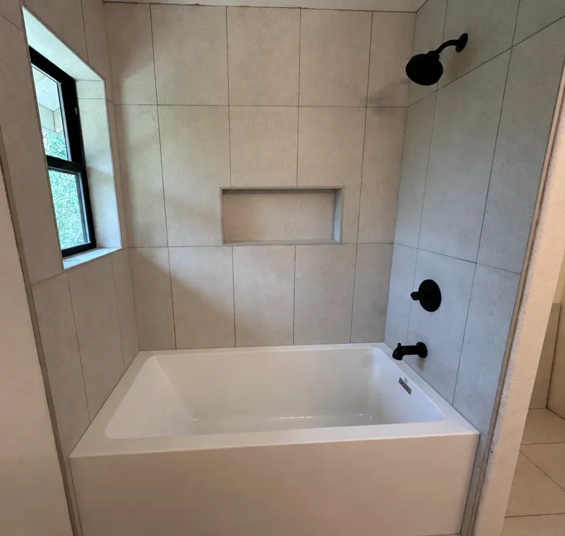 A white tub beneath light beige-gray rectangular wall tile, with black shower fixtures, a recessed niche and a dark-framed window.
