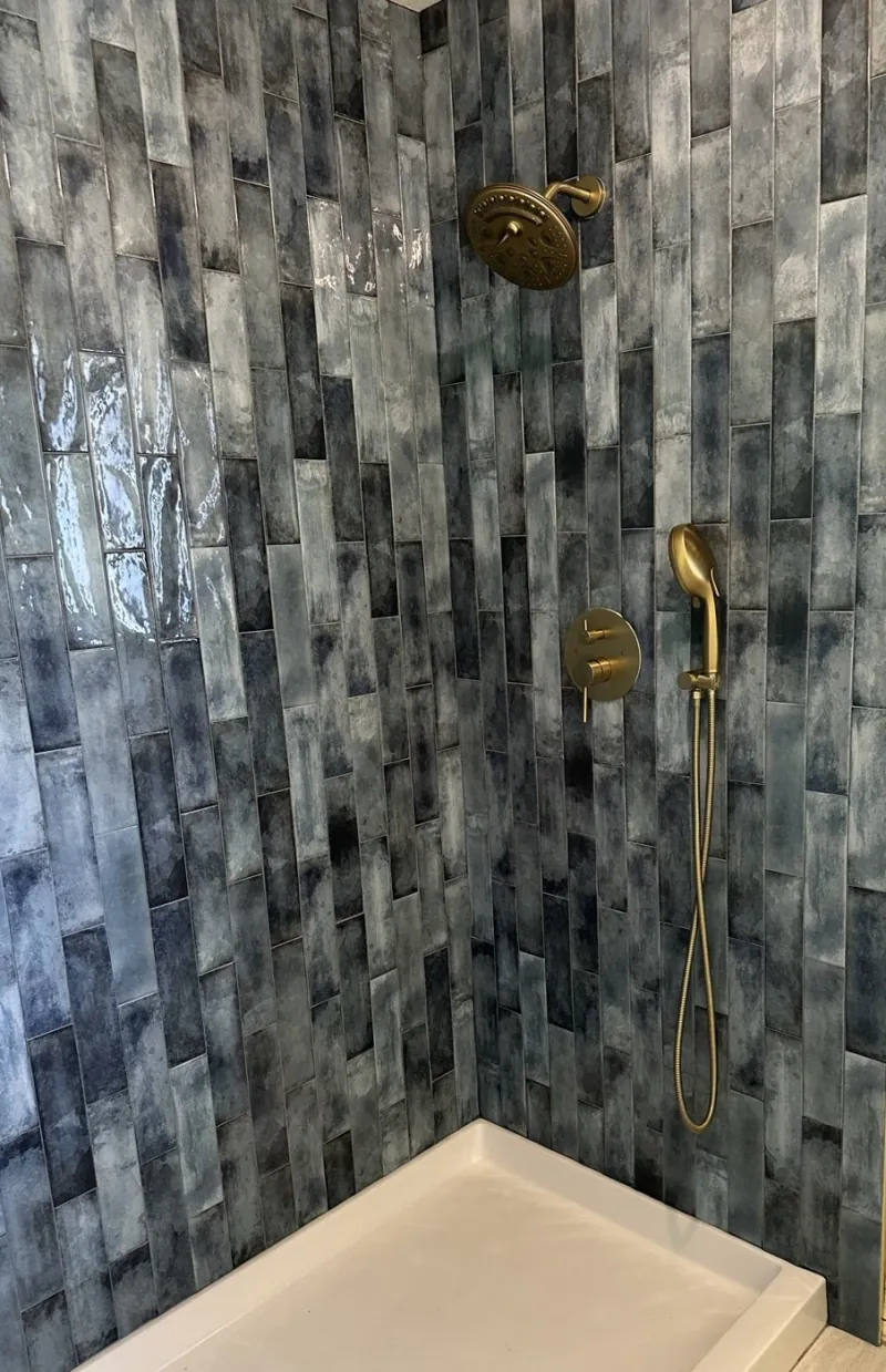 Glossy blue-gray rectangular tiles run vertically around warm-metal shower fixtures above a white shower tray.