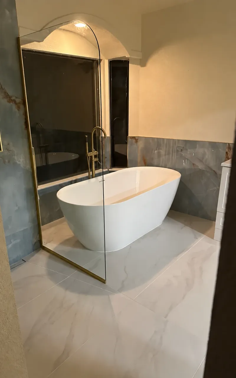 A white freestanding tub beside blue-green veined walls, pale veined floor tile and glass shower panels.