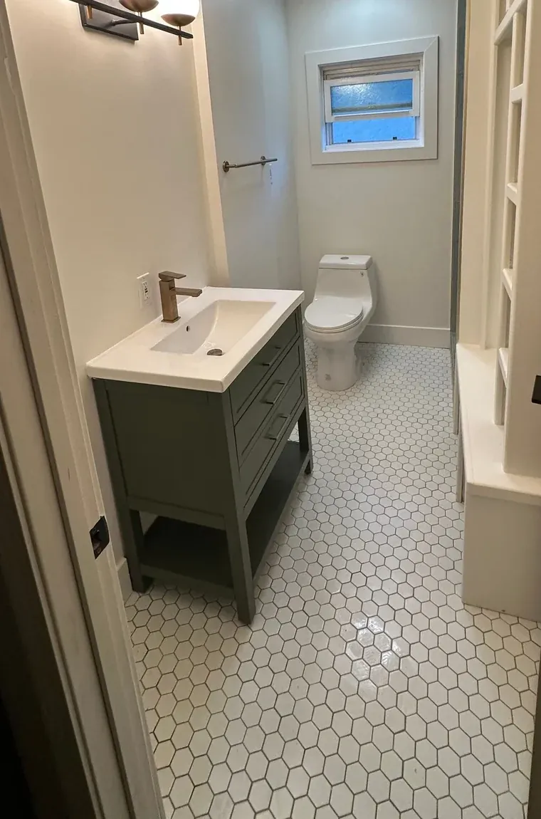 White hexagonal floor tiles with dark grout run between a gray vanity and a toilet beneath a small window.