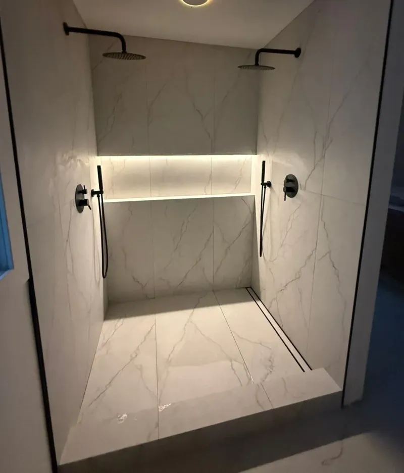 Two black overhead showerheads and handheld sprayers flank an illuminated niche above pale veined tile and two linear floor drains.