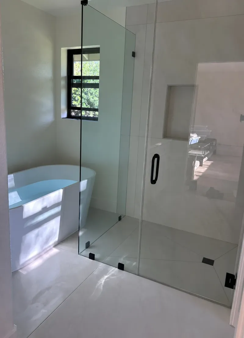A white freestanding tub beside a clear-glass shower with pale wall and floor tile, a recessed niche and black hardware.