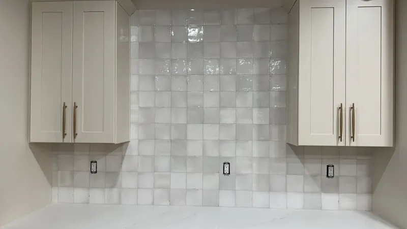 Glossy white square tiles form a straight grid backsplash between white kitchen cabinets above a pale counter.