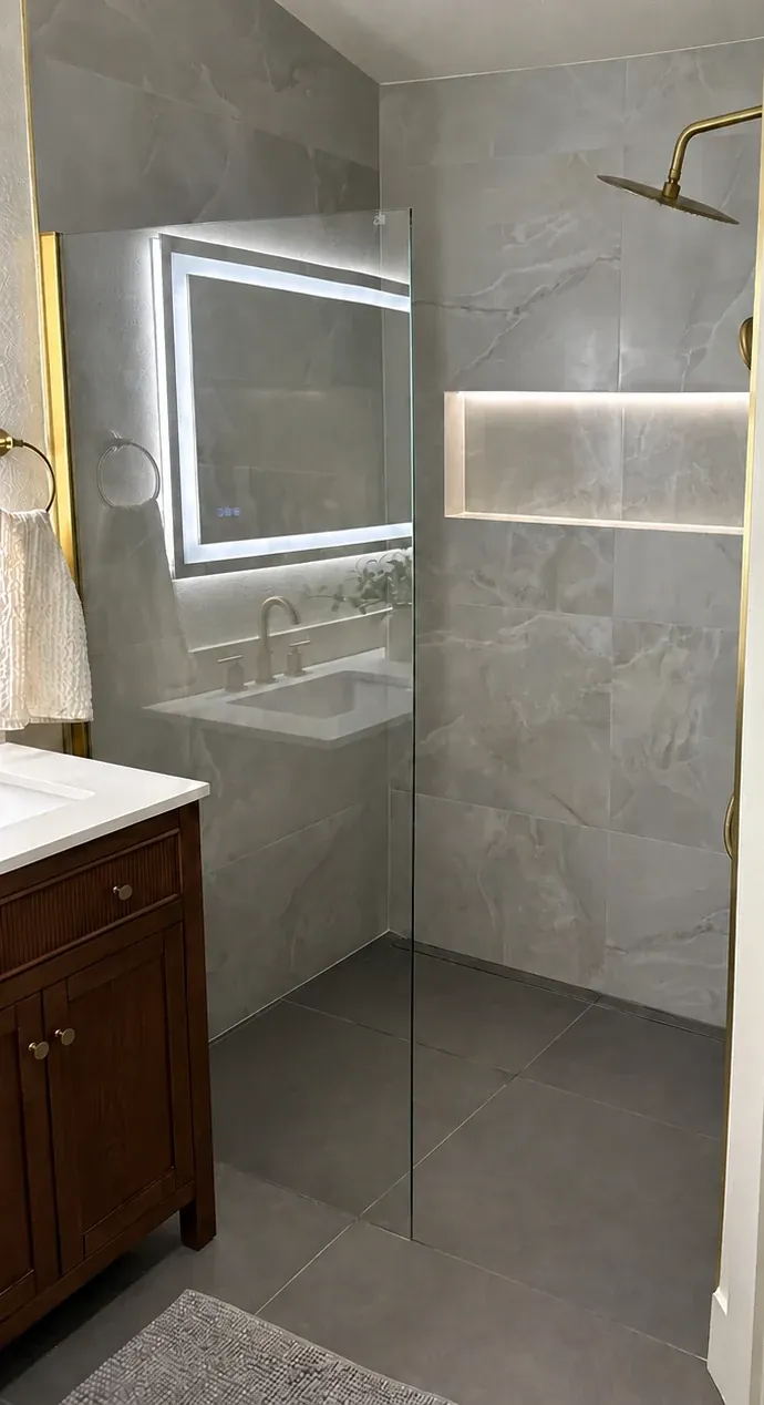 A clear-glass shower with gray-veined wall tile and a lit recessed niche beside a dark wood vanity and illuminated mirror.