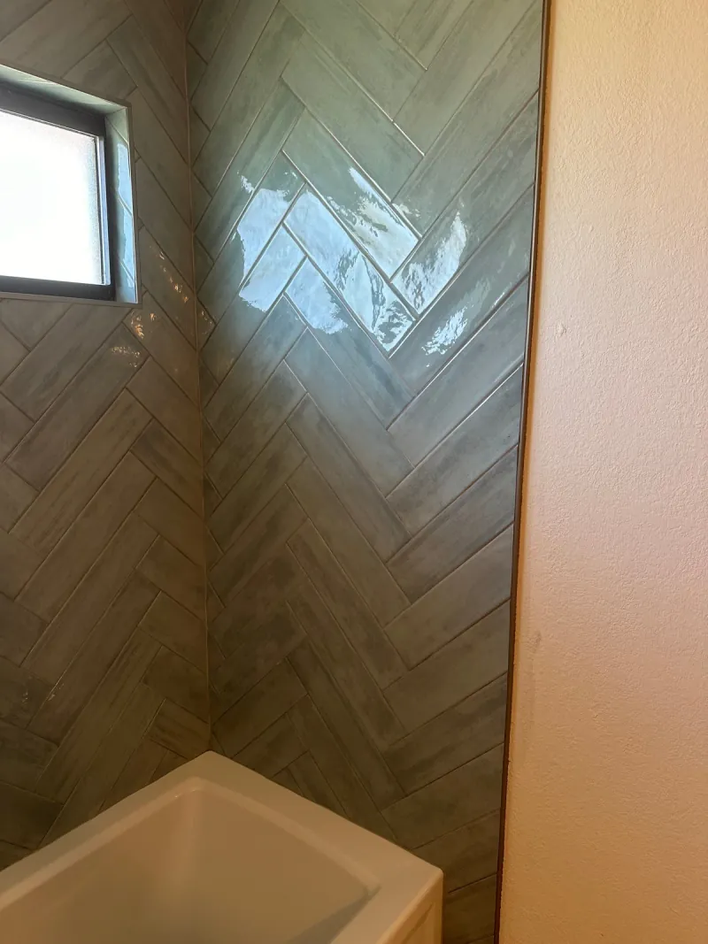 Glossy blue-gray rectangular tiles form a herringbone pattern above a white tub and around a frosted window.