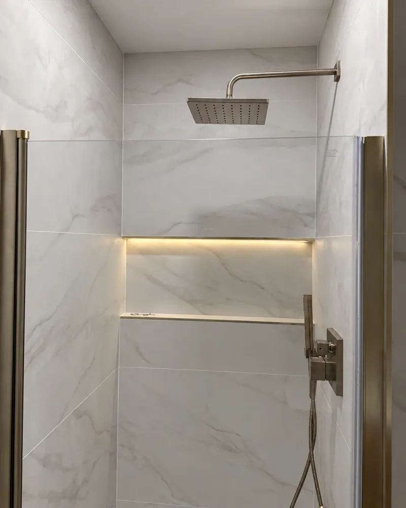 A long recessed niche illuminated along its top edge in pale veined shower tile beneath a square showerhead, with warm-metal fixtures beside the glass screen.