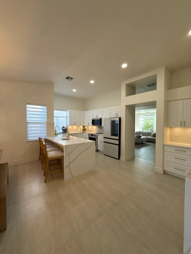 Pale rectangular flooring extends around a veined peninsula and white kitchen cabinetry, with an adjoining living area beyond the doorway.