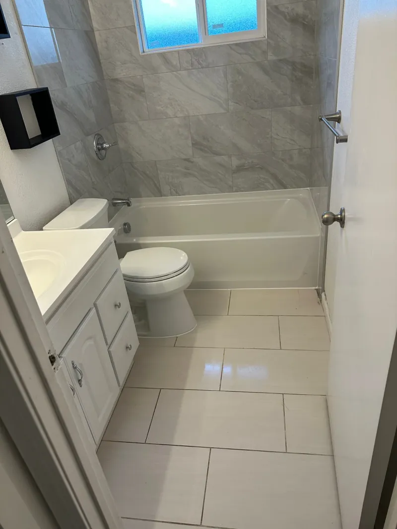 A white tub sits beneath gray-veined rectangular wall tile, with pale rectangular floor tile, a white vanity and a toilet.