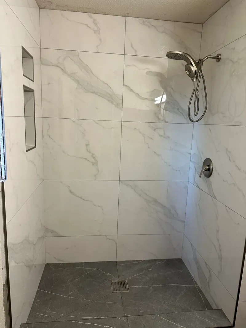 Gray-veined light wall tile, two recessed niches and a dark tiled floor inside a shower with a handheld shower fixture and square drain.