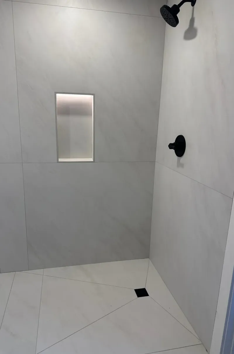 Light gray-veined wall and floor tile in a shower with black fixtures, a lit recessed niche and a square floor drain.