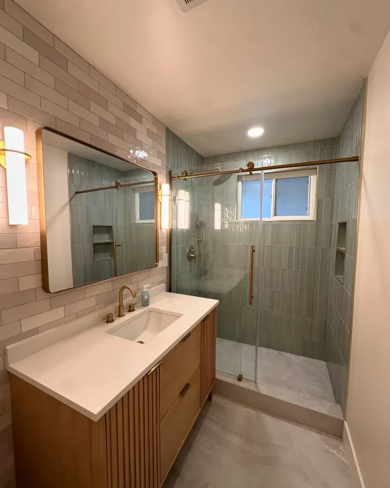 A wood double vanity with white basins faces a glass shower lined with sage-green vertical tile, brass framing and fixtures, with white subway tile around the mirrors.