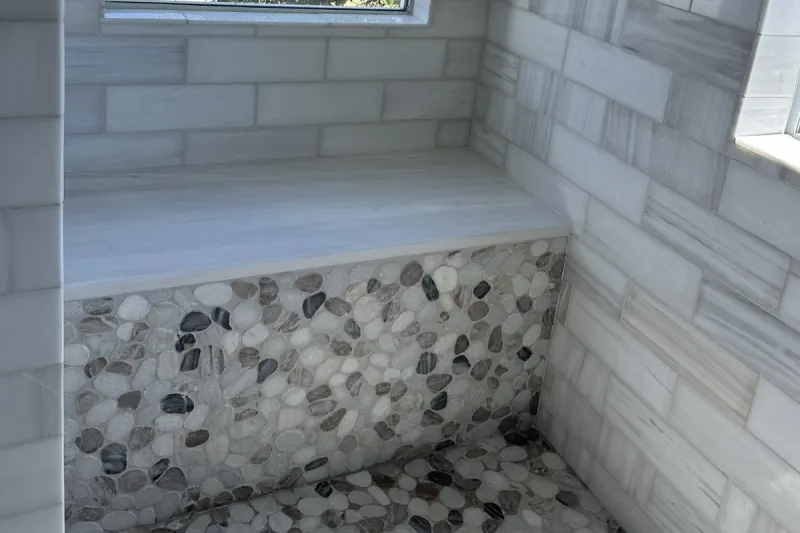 A tiled shower bench beneath a window has a pebble mosaic front beside pale rectangular wall tile and pebble floor mosaic.