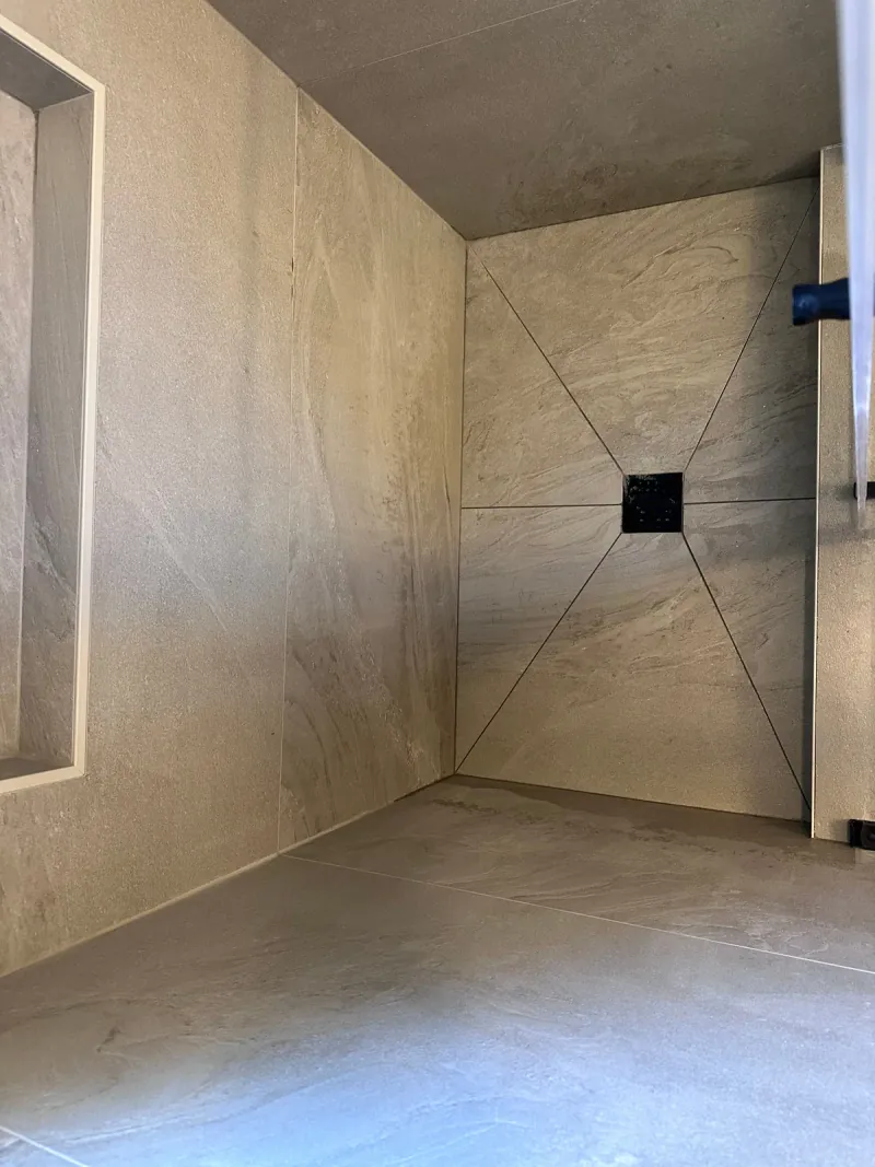 Large beige veined shower floor tiles meet a black square drain, with diagonal joints forming an envelope-cut layout.