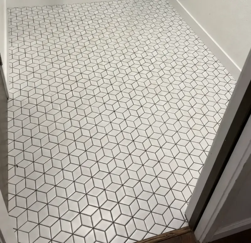 White angular mosaic pieces and dark grout form a cube-like pattern across a floor beside white baseboards and a wood threshold.