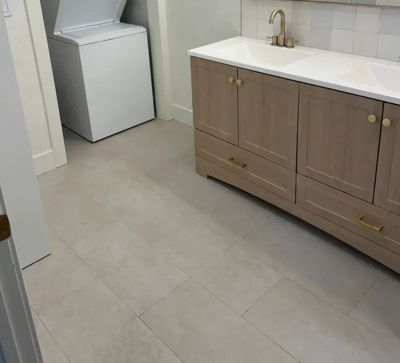 A close view of pale rectangular bathroom floor tiles beside a wood vanity and a white laundry appliance.