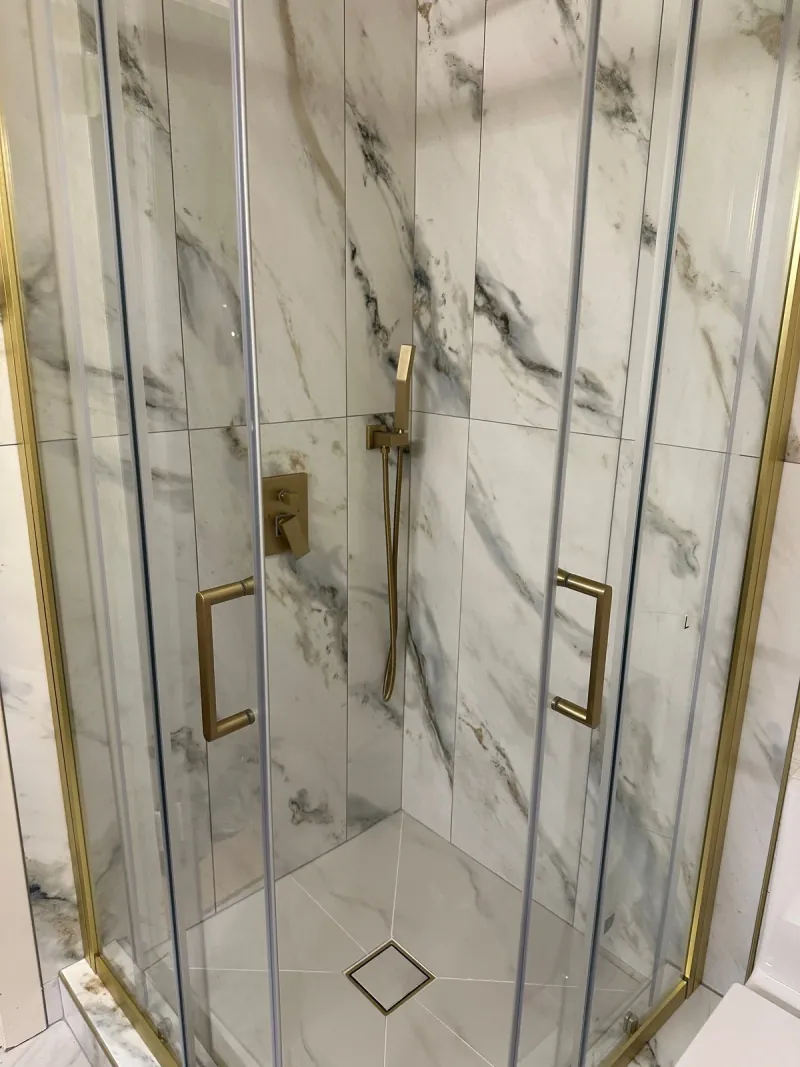 Tall rectangular white tile with gray veining lines the walls of a glass corner shower with warm-metal framing and a square floor drain.