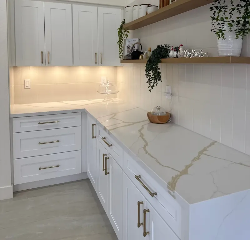 A white counter with gold-gray veining meets a pale backsplash of narrow vertical rectangular tiles beneath open wood shelves.