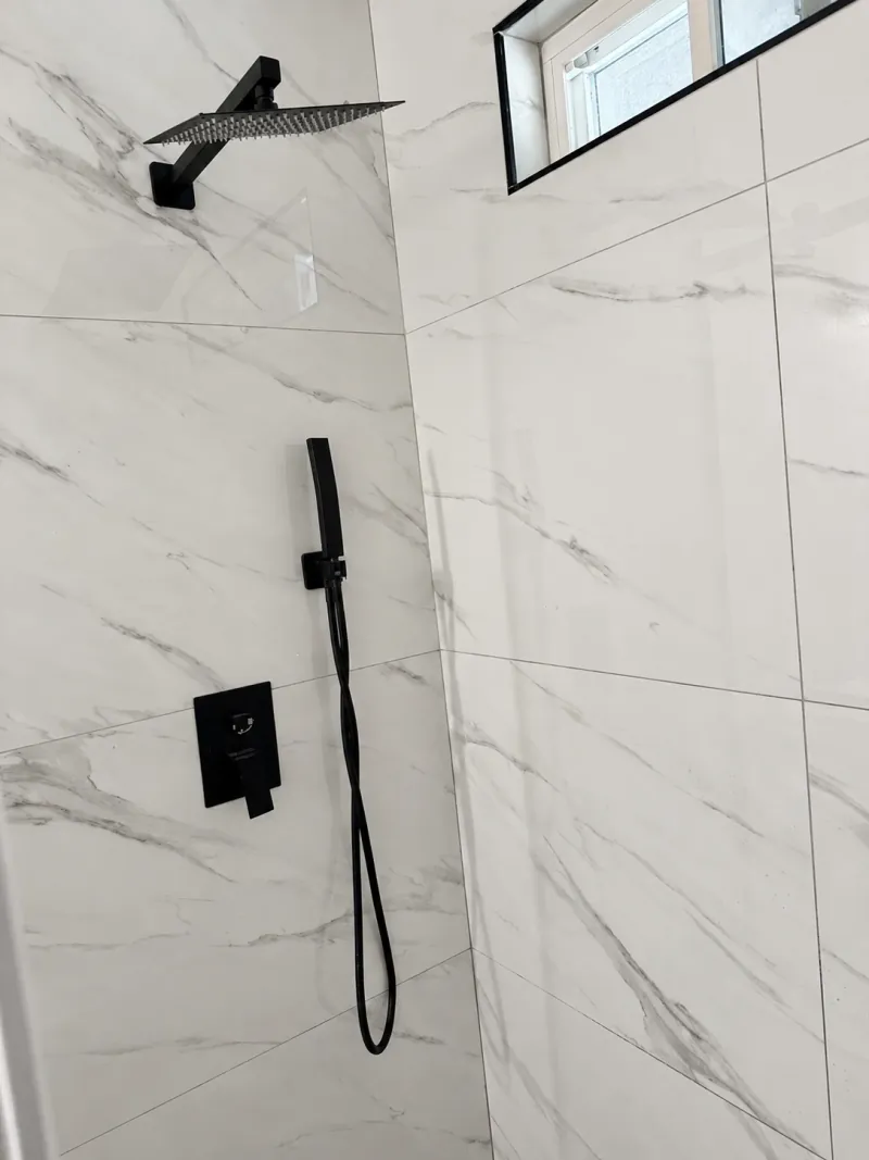 Pale gray-veined rectangular shower tiles meet at a corner beneath a high window, with a black overhead showerhead, handheld sprayer and controls.