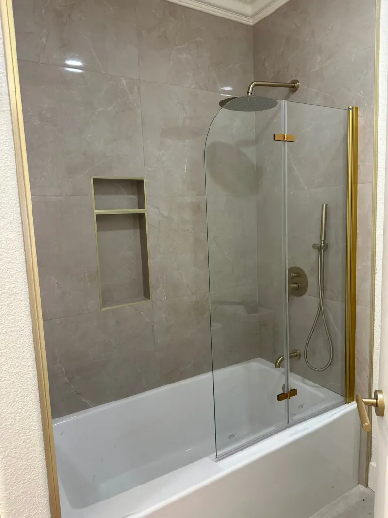 Gray-beige veined rectangular tile around a white tub, with recessed storage, a glass screen and gold-colored shower fixtures.