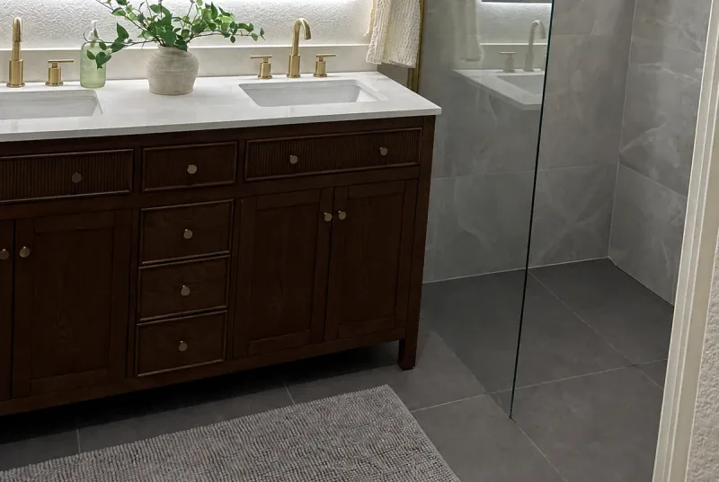 Large gray floor and shower wall tiles sit beside a dark wood double vanity with a white top and glass shower enclosure.