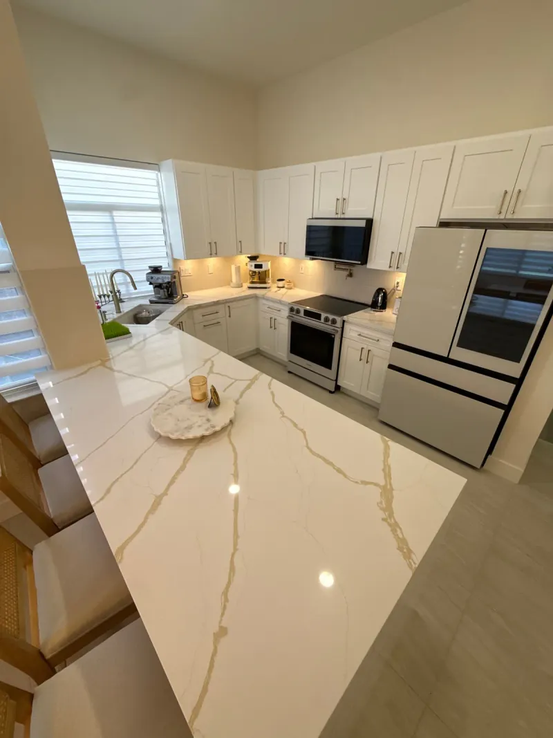 A pale veined peninsula faces white kitchen cabinetry, a sink, range and refrigerator, with rectangular flooring below.