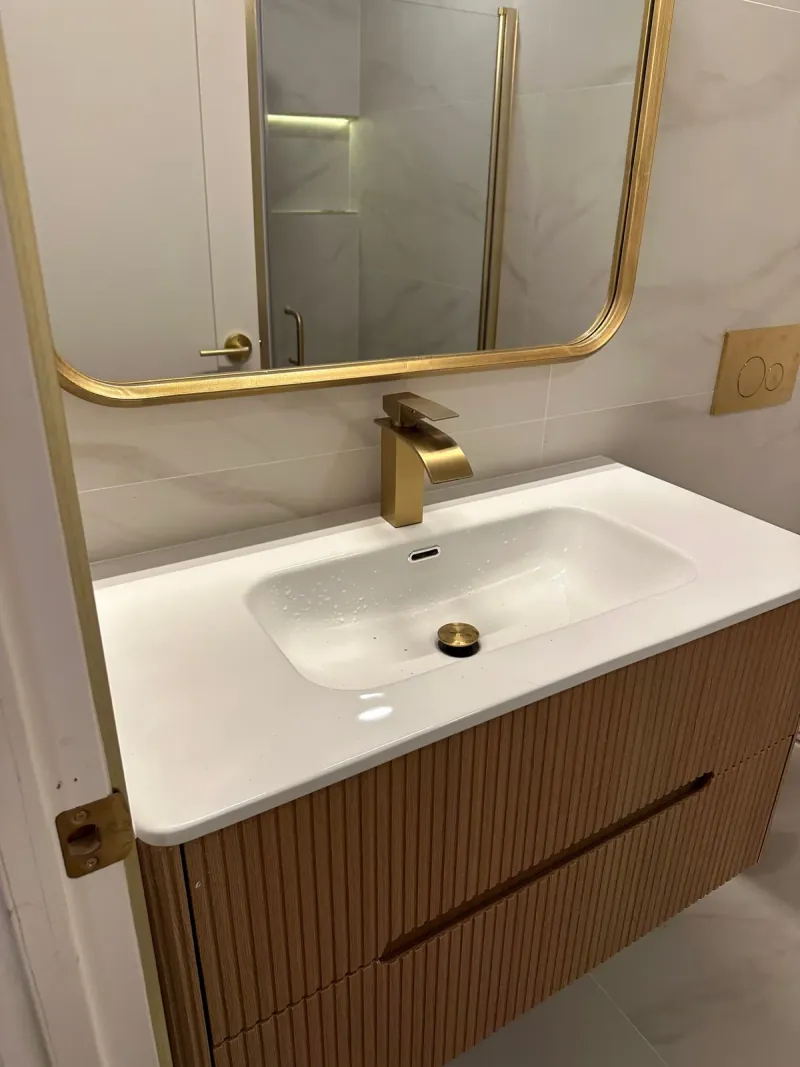 A white vanity top with an inset basin sits beneath a large warm-metal framed mirror, against pale veined wall tile and fluted wood drawers.