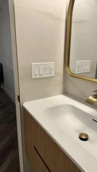 White basin and angular warm-metal tap beside pale veined wall tile and a mirror.