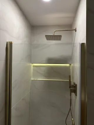 A long illuminated niche in pale veined shower tile below a square warm-metal shower head.