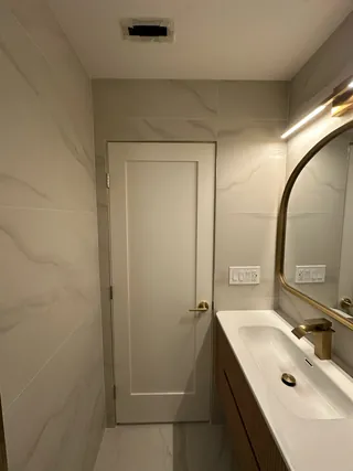 Pale veined wall and floor tile beside a fluted wood-tone vanity and closed bathroom door.