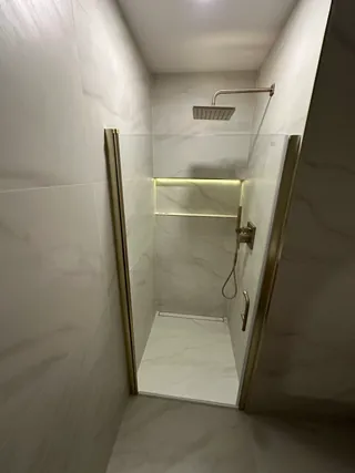 Pale veined shower tile behind clear glass, with warm-metal fixtures, a lit niche and a linear floor drain.
