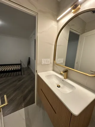 A white basin and fluted wood-tone vanity beneath a rounded warm-metal mirror beside an open doorway.