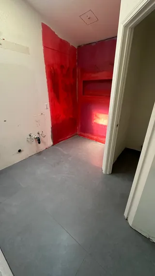 Gray rectangular bathroom floor tile in place beside red-coated shower walls during installation.