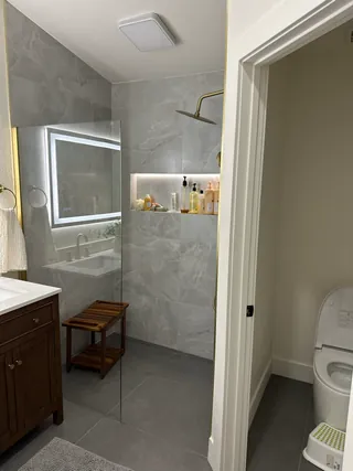 Clear-glass shower with pale veined wall tile, a long illuminated niche, gold-tone fittings and a loose wooden stool.