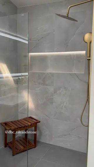 Gold-tone shower fittings beside a long illuminated niche in pale veined wall tile, with clear glass and a wooden stool.