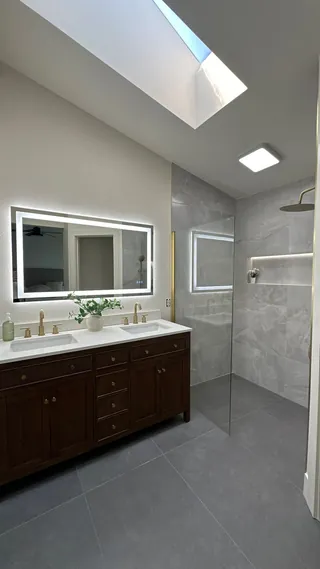 Skylight above a dark wood-toned double vanity, gray tile floor and clear-glass shower with a long illuminated niche.