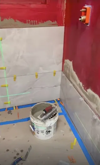 Pale veined shower wall tile being set beneath red-coated wall areas, with tile clips, a visible laser line and taped floor protection.