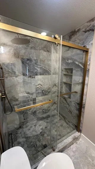 Gold-tone sliding glass shower hardware in front of gray-veined wall tile and a hexagonal mosaic floor.