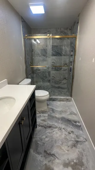 Gray-veined tile across a bathroom floor and glass shower beside a dark vanity with gold-tone hardware.