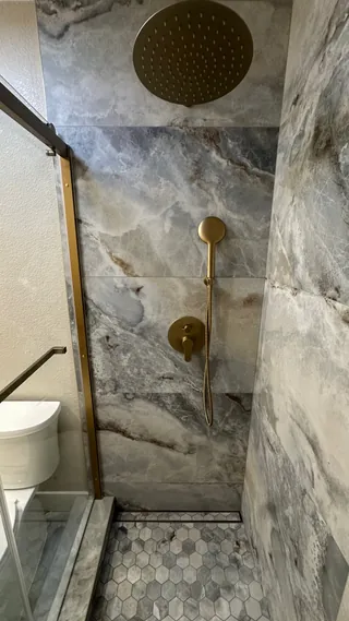 Round gold-tone showerhead and handheld spray against gray-veined wall tile above a hexagonal mosaic floor.
