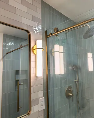 Gray offset wall tile meets vertical sage shower tile beside a mirror, wall light and gold-tone glass rail.
