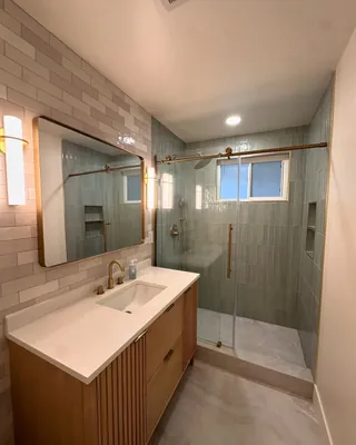 Sage-green vertical shower tile and gray offset wall tile beside a fluted wood-tone vanity, with clear glass and gold-tone fittings.