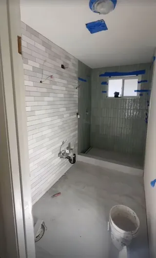 Gray offset wall tile and vertical sage shower tile installed in an empty bathroom, with taped window edges and unfinished fittings.