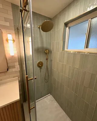 Gold-tone overhead and handheld shower fittings against vertical sage tile beside a frosted window and clear glass.