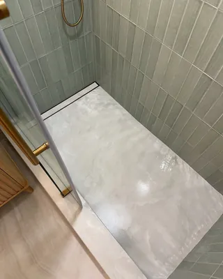 Pale shower floor with a long narrow drain opening beside sage tile walls and a clear-glass threshold.