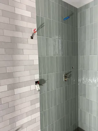 Vertical sage shower tile meets gray offset wall tile, with unfinished fixture openings and exposed electrical fittings.