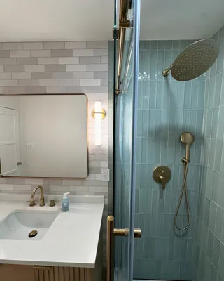 Wood-tone vanity, gray offset wall tile, mirror and gold-tone shower fittings seen across a clear-glass panel.