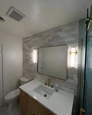 Gray and white offset rectangular wall tile behind a mirror and fluted wood-tone vanity, with a toilet at one side.