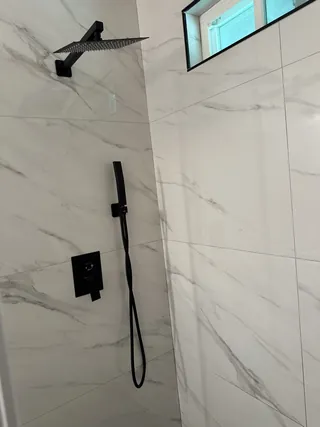Square black overhead showerhead and handheld spray against white veined tile beside a high window.