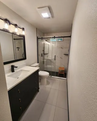 Bathroom with a dark vanity, pale rectangular floor tile and white veined shower tile beneath a high window, behind black sliding glass hardware.