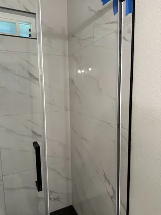 Clear shower glass and a black handle in front of white tile with gray veining.